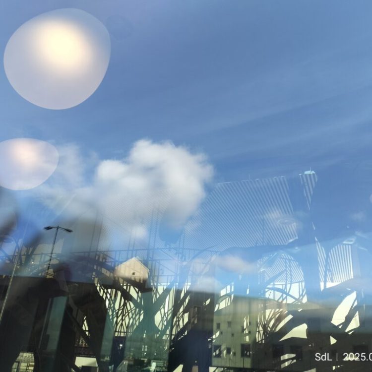 Wolken in een blauwe lucht weerspiegeld door grote ramen van een modern gebouw. Foto: Sacha de Leeuw